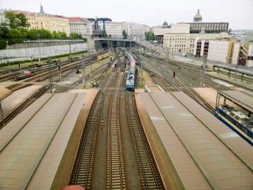 Železniční nádraží s vlakem přijíždějícím k perónu, obklopené městskou architekturou a kolejemi.